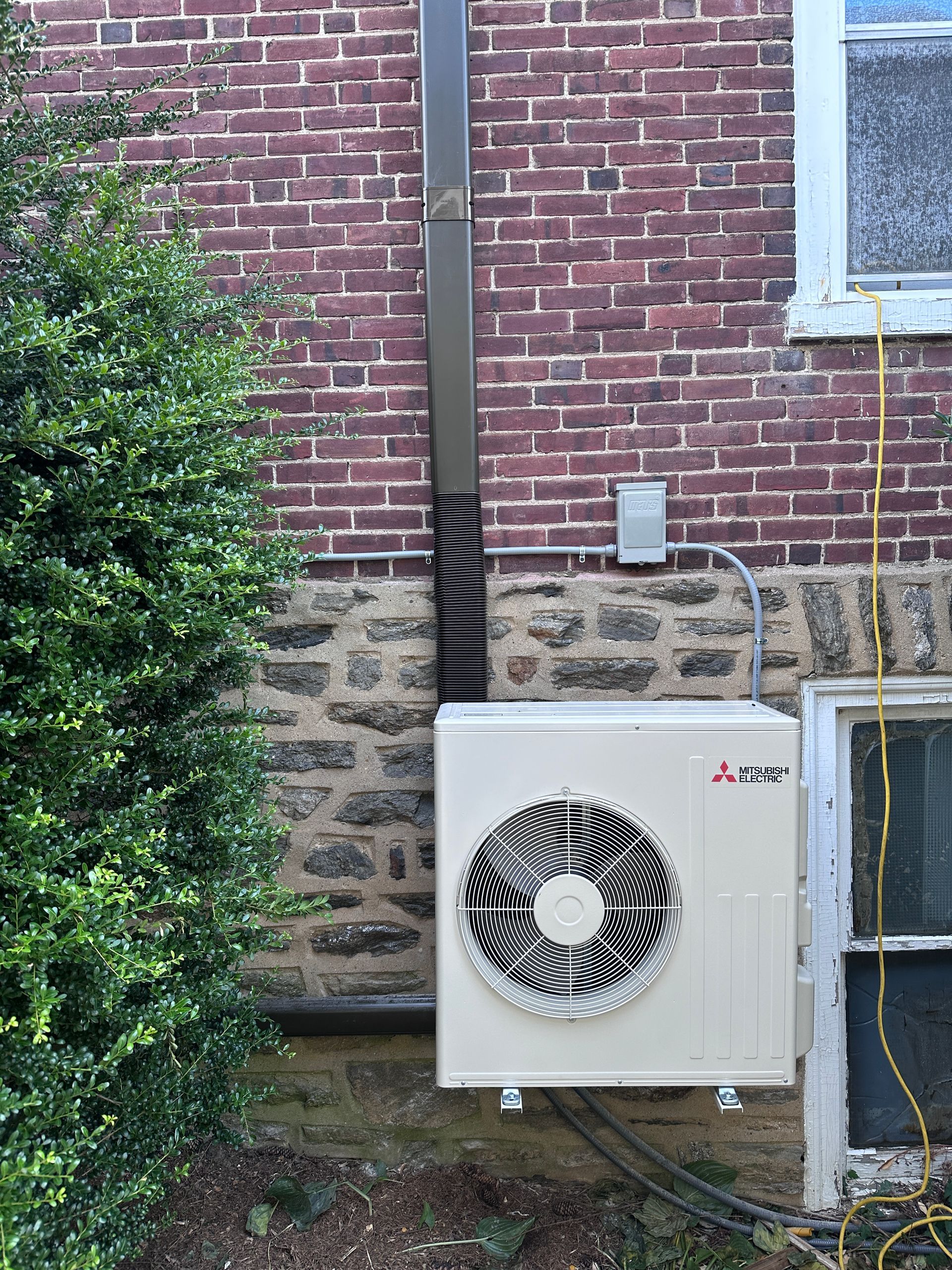Mitsubishi heat pump unit mounted on brick wall, next to a bush and window.