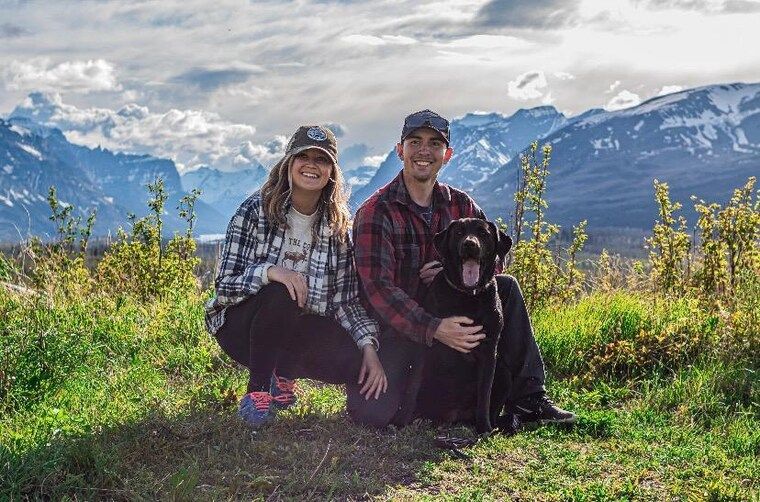Two people kneel with a black dog in a mountain meadow, smiling under cloudy skies.