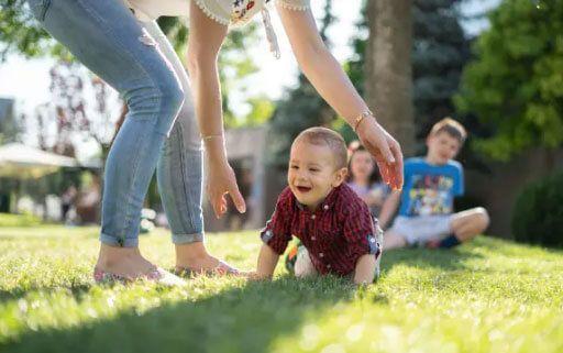 A woman is helping a baby crawl in the grass.