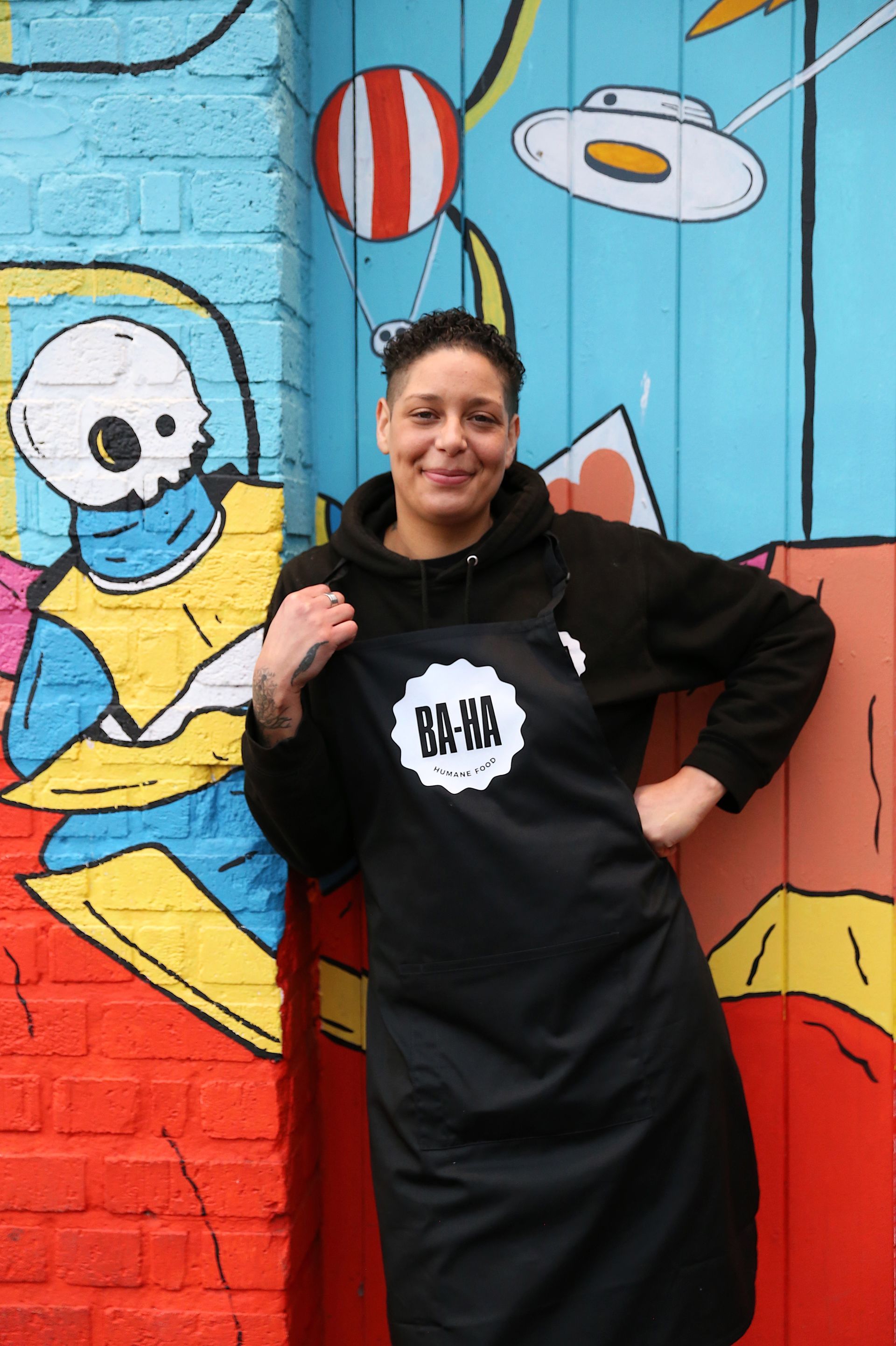 Woman in black apron with logo, smiling next to colorful mural.