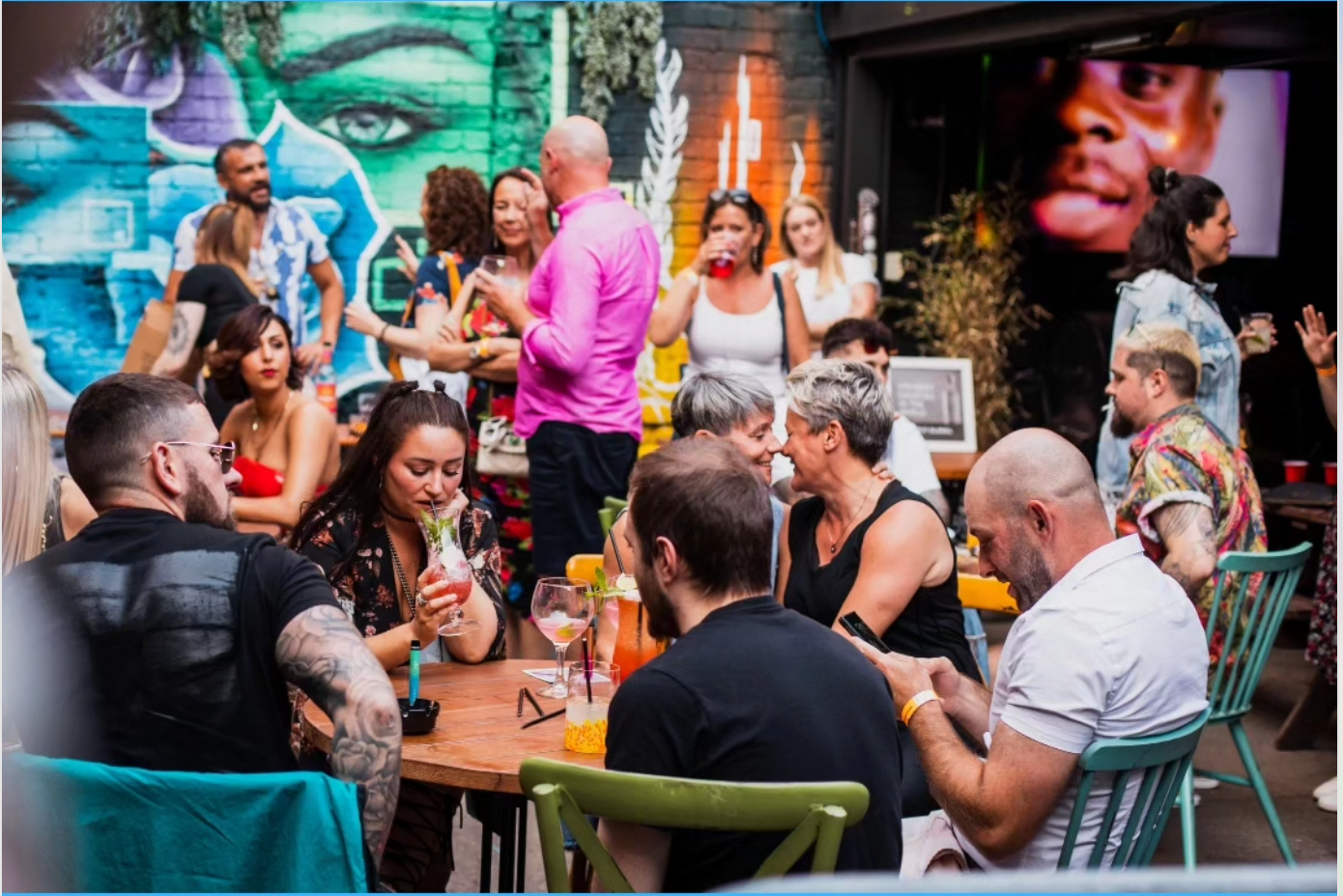 People socializing outdoors, drinking and talking at tables. Graffiti mural in background.