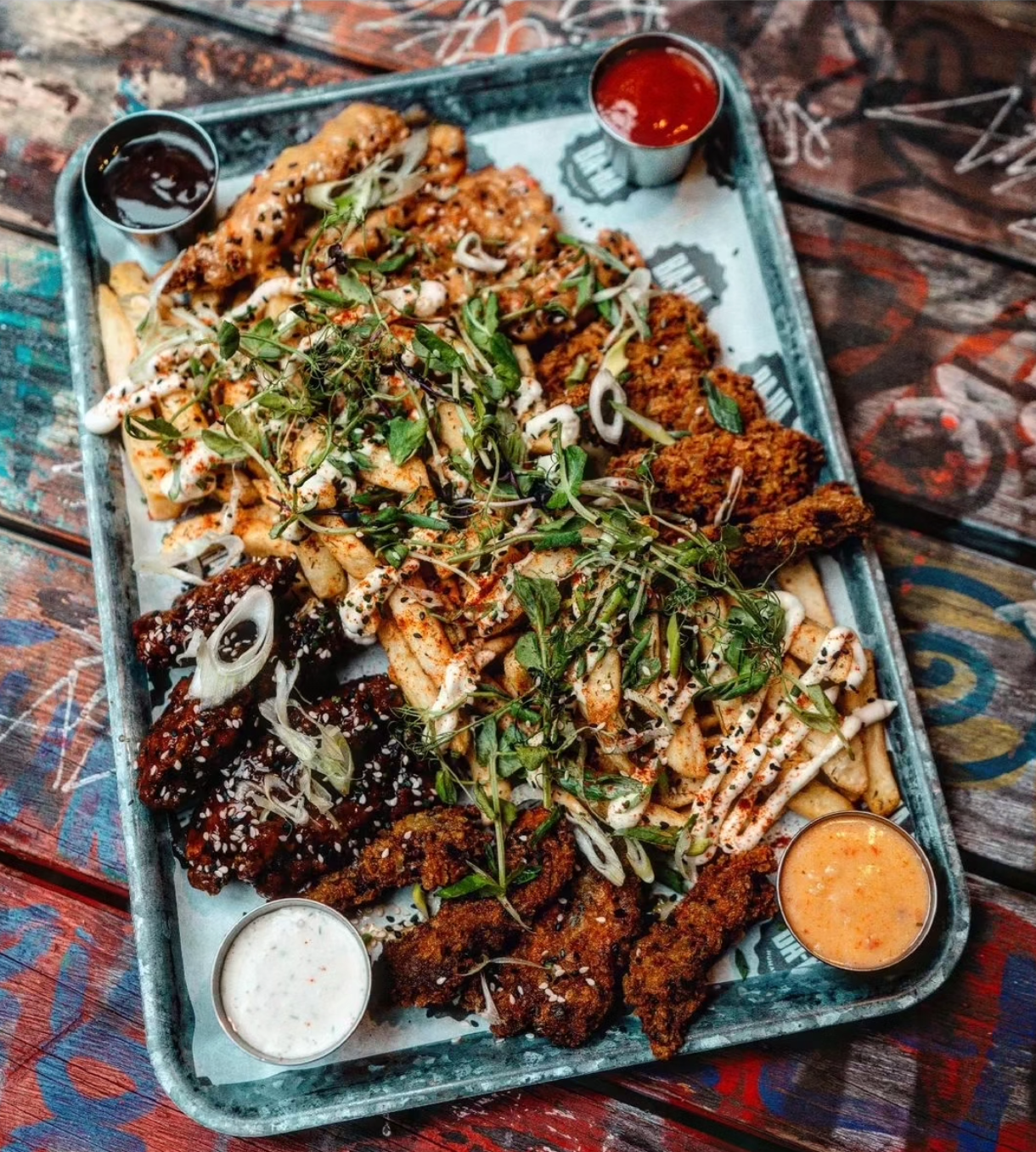 Large tray of fried chicken and fries with dipping sauces, garnished with greens, on a graffiti-covered table.