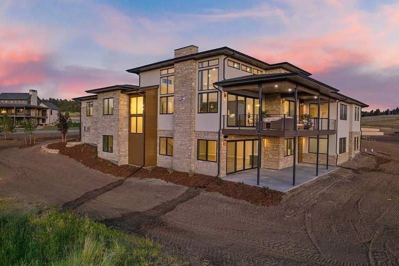 Modern two-story house at dusk with lit windows and a covered patio in a rocky suburban setting