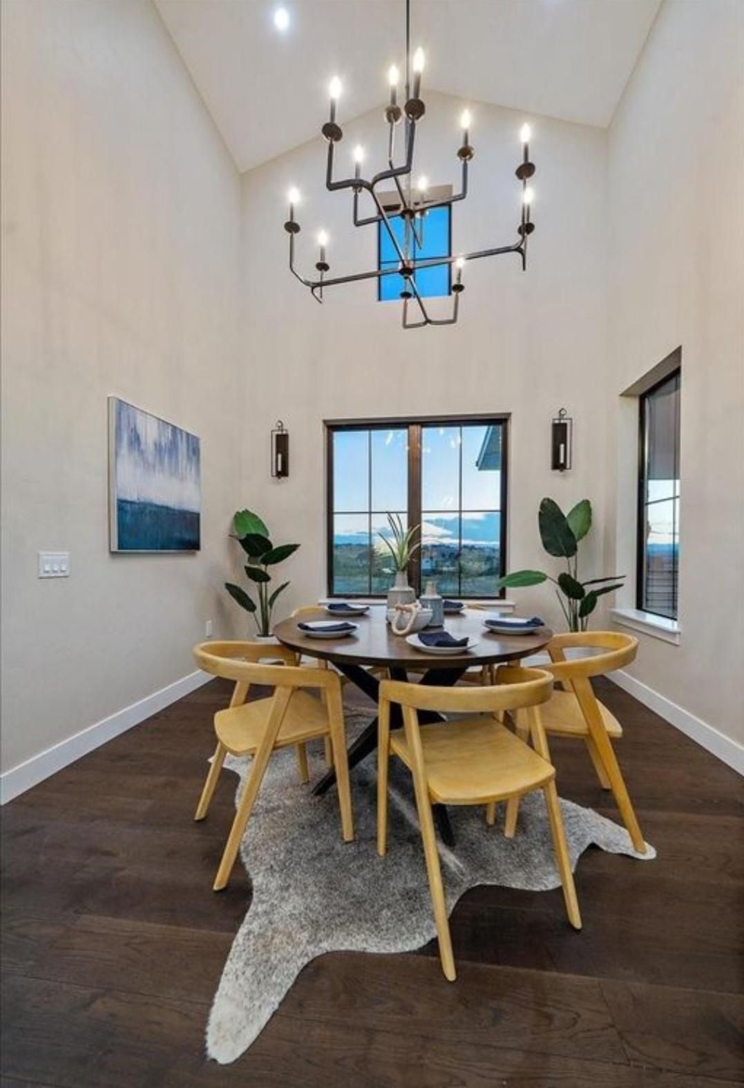 Modern dining room with round wooden table, six chairs, chandelier, and large windows overlooking the outdoors