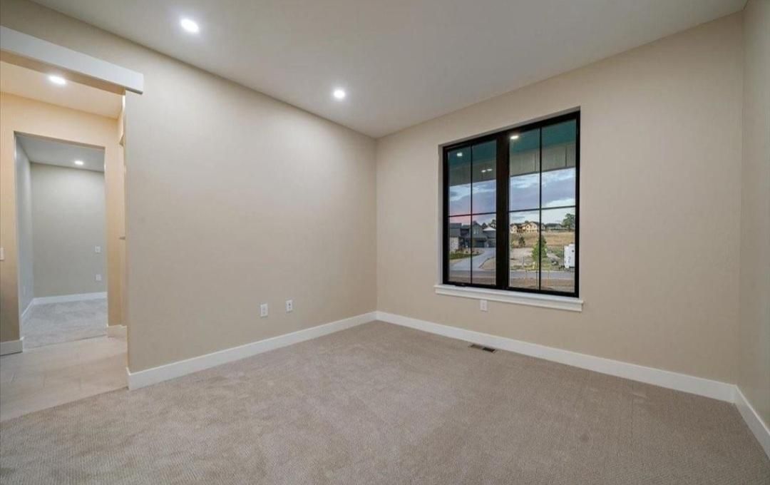 Empty beige bedroom with carpet, recessed lights, and a window overlooking the street.