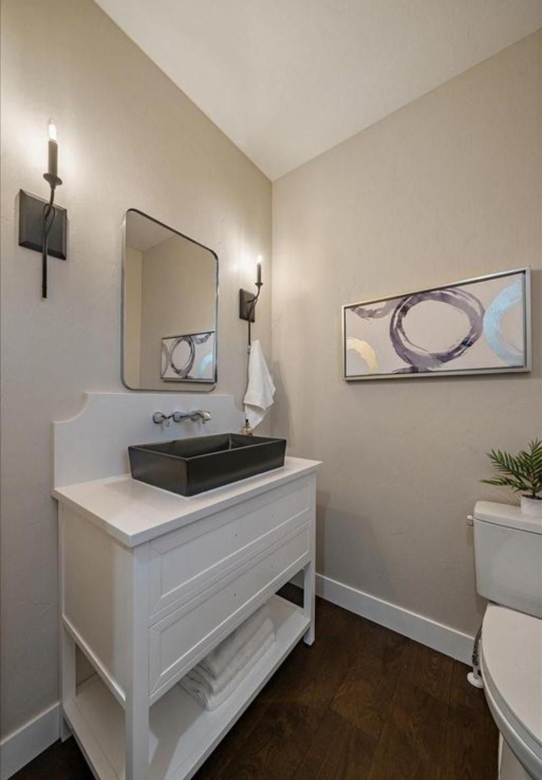 Small bathroom vanity with black sink, wall mirror, sconces, and abstract art on beige walls.