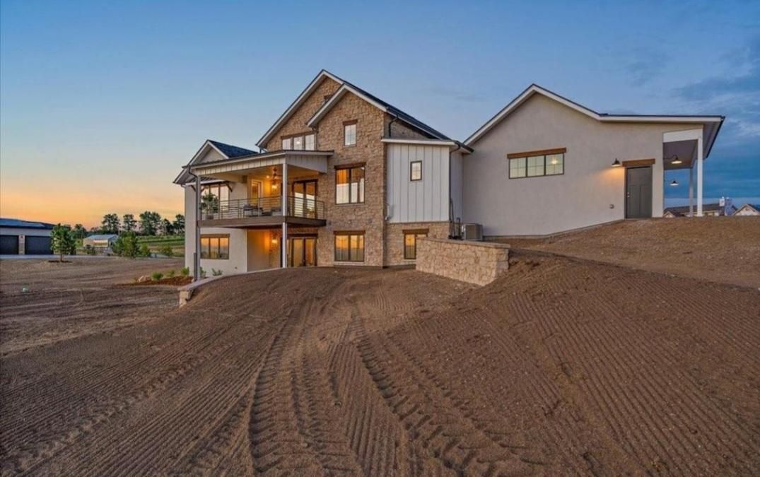 Large modern house under construction at dusk with lit balconies and a dirt lot in front