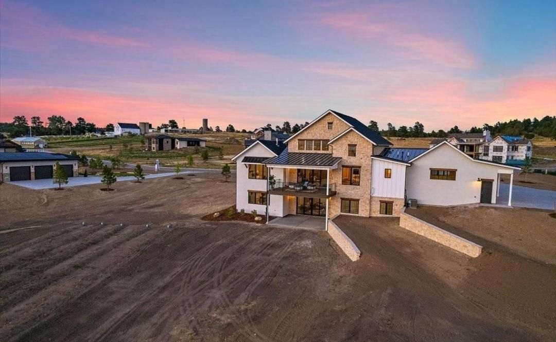 Large white farmhouse under pink sunset sky with open dirt foreground and neighboring houses nearby