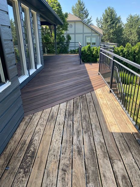 Weathered wooden deck beside a house, with black railing, glass doors, and a sunlit yard beyond.