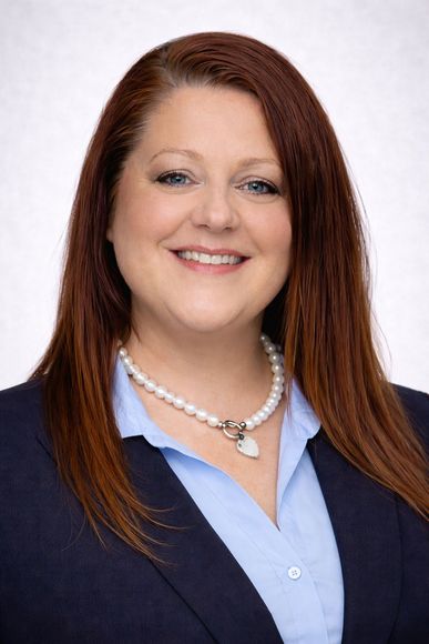 Headshot of a smiling woman with long brown hair, wearing a navy blazer and light blue shirt against a white background