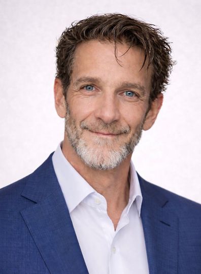 Studio headshot of a smiling man in a blue suit jacket and white shirt against a light background