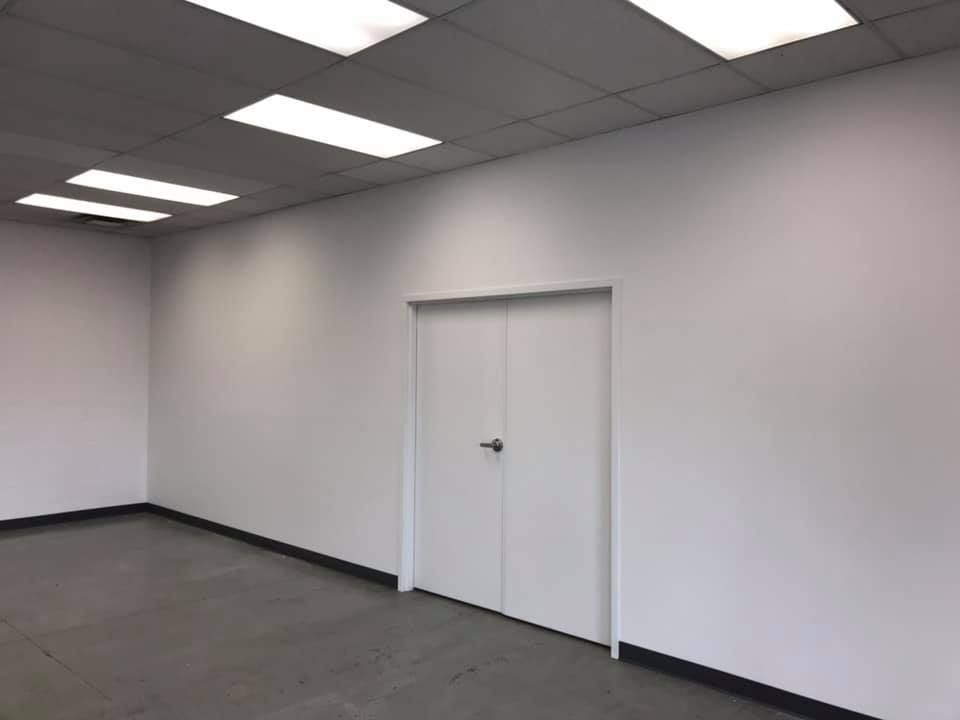 Empty office room with gray carpet, white walls, ceiling lights, and a closed white door