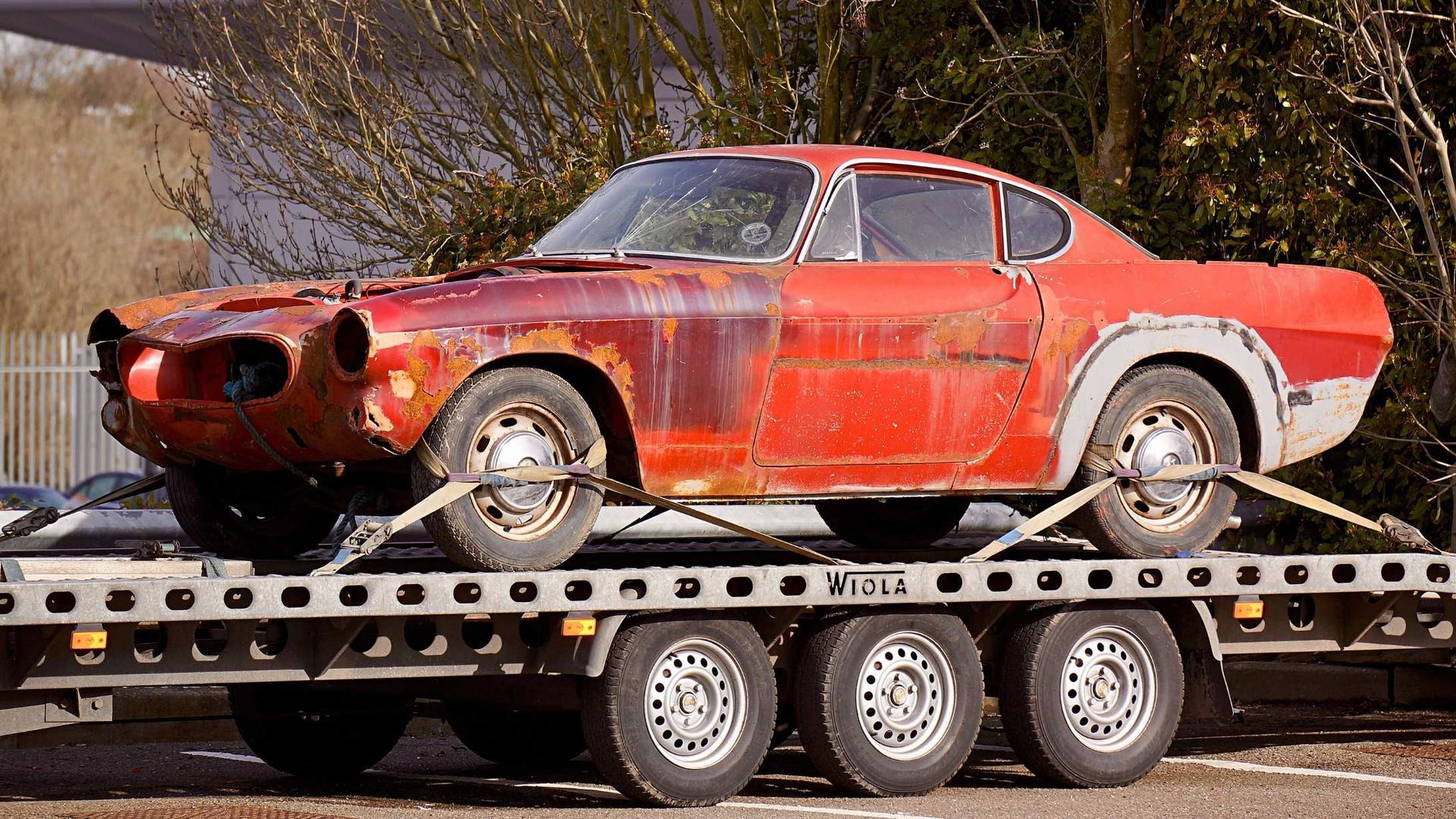Rusty red vintage car strapped on a flatbed trailer, missing front parts, outdoors.