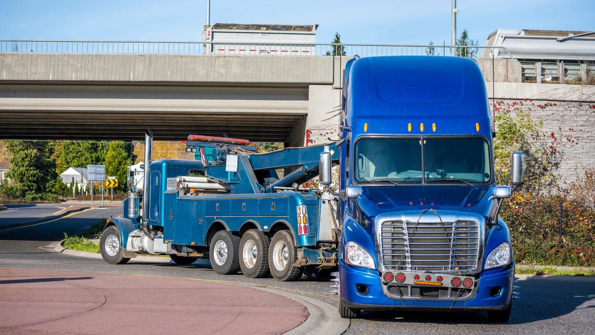 Blue semi-truck and tow truck parked under an overpass on a city street.