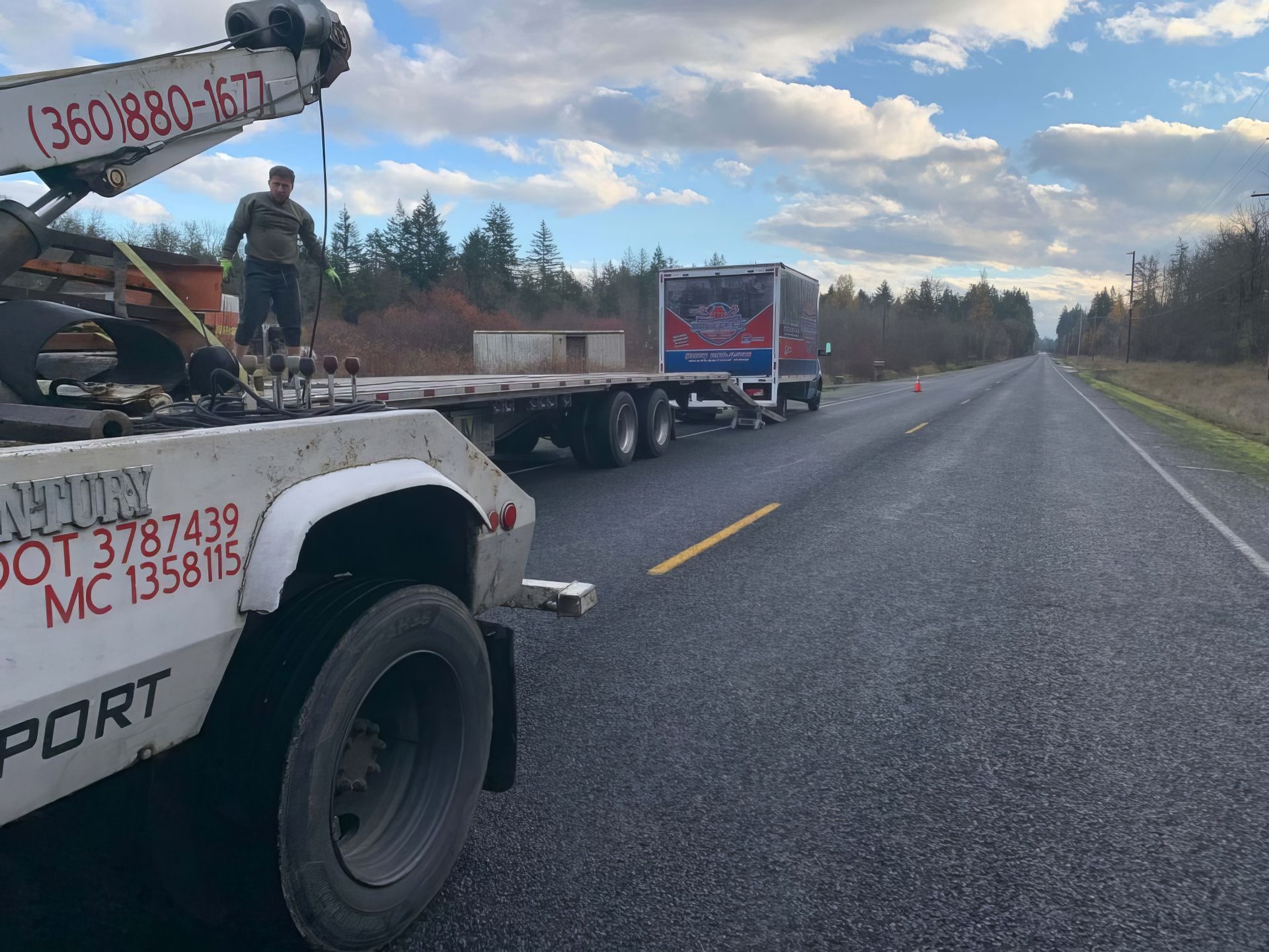 Tow truck and flatbed with car on roadside shoulder under cloudy sky