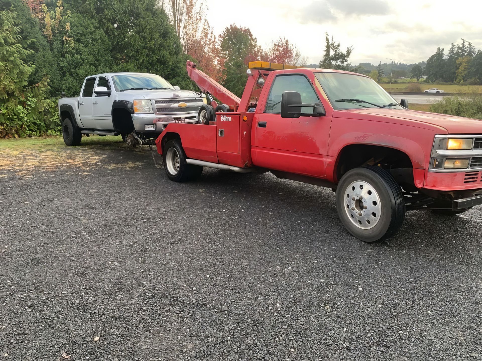 Red tow truck pulling a white pickup on a gravel road at dusk