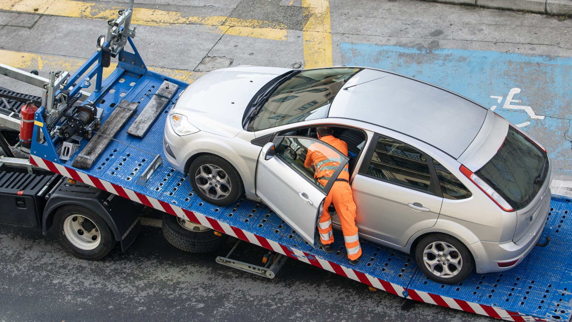 Tow truck worker in orange beside silver car on blue flatbed tow truck on city street
