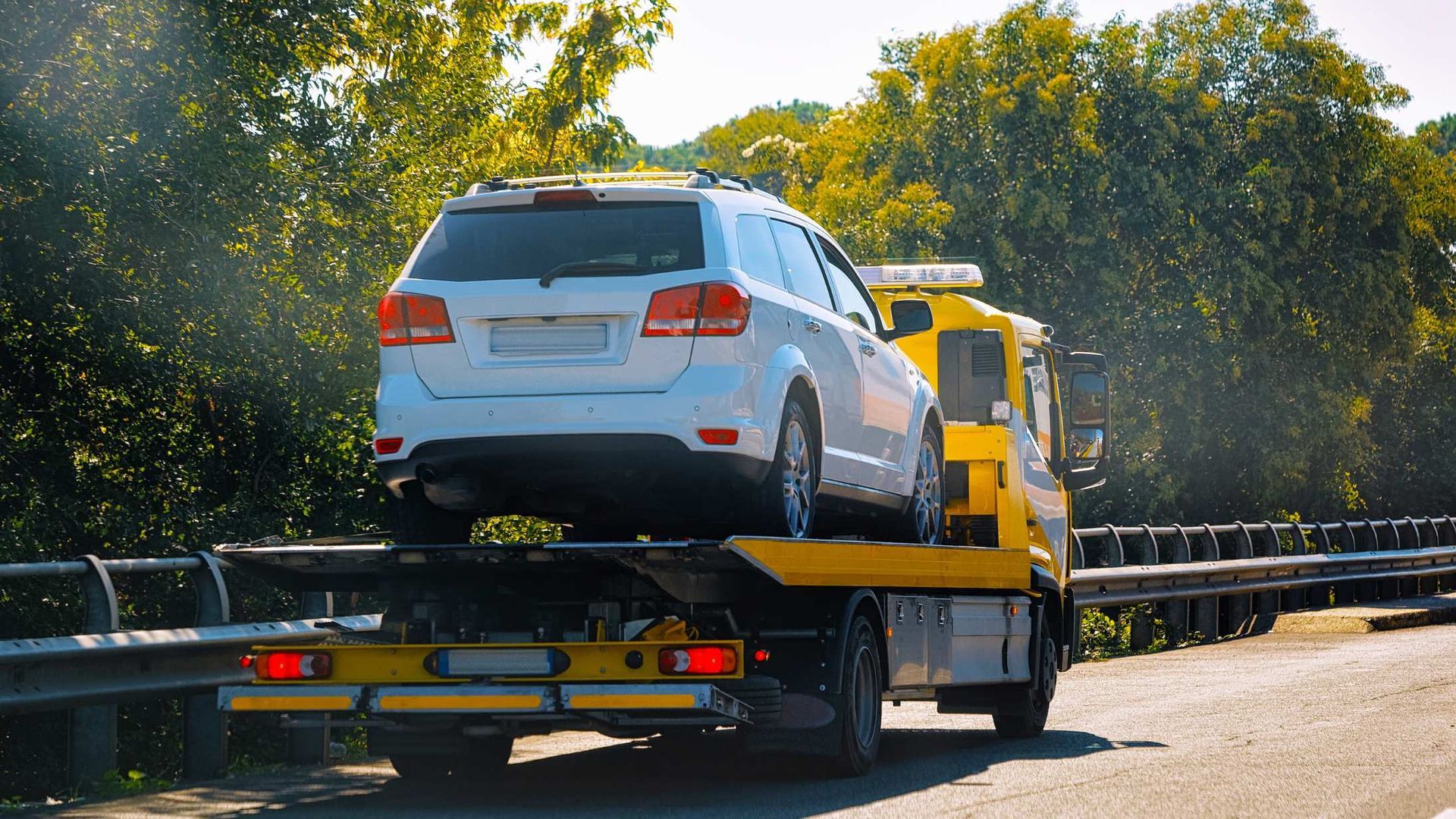 White SUV loaded on a yellow tow truck driving on a sunny road beside guardrails and trees.