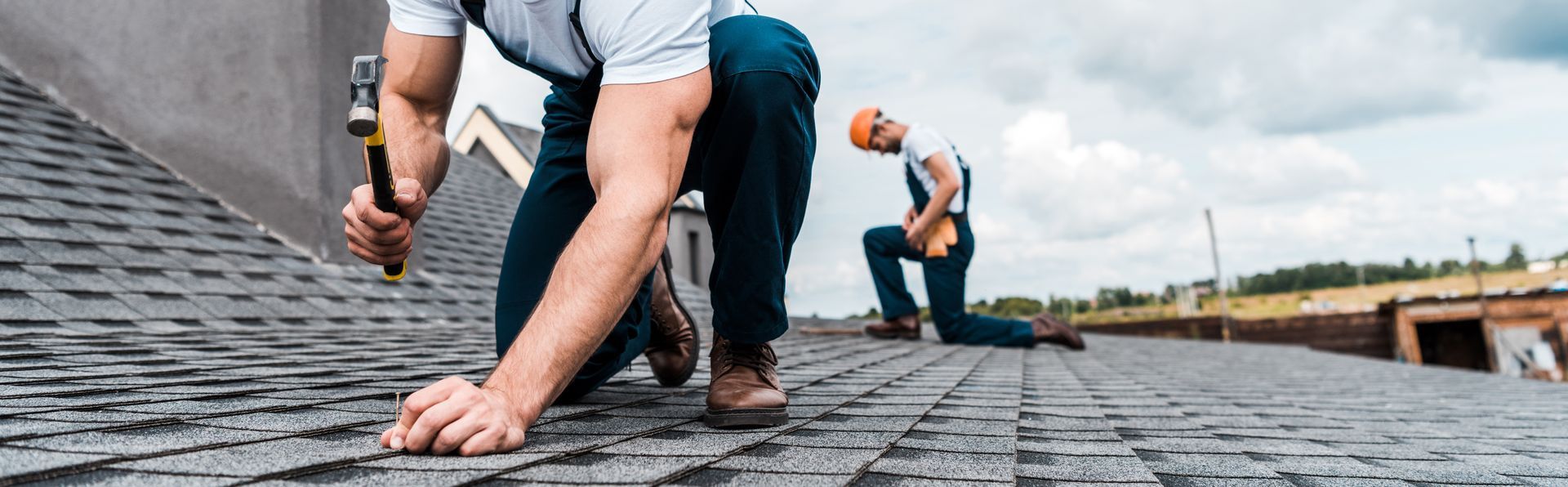 Two roofers install shingles, one hammering nails while another works in the background. Two roofers install shingles, one hammering nails while another works in the background.