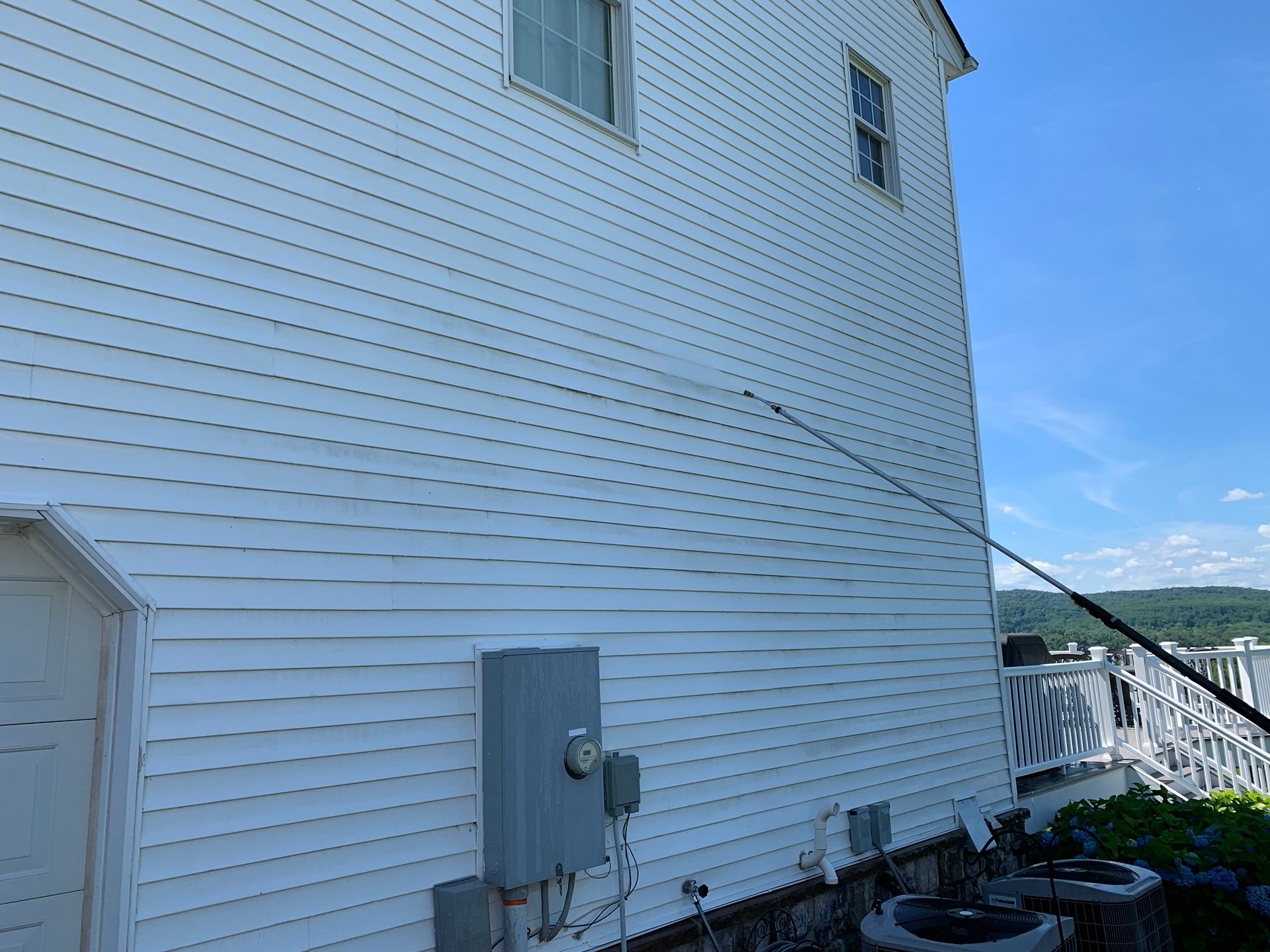 A power washer nozzle sprays water onto white vinyl siding on the exterior of a house under a clear blue sky.