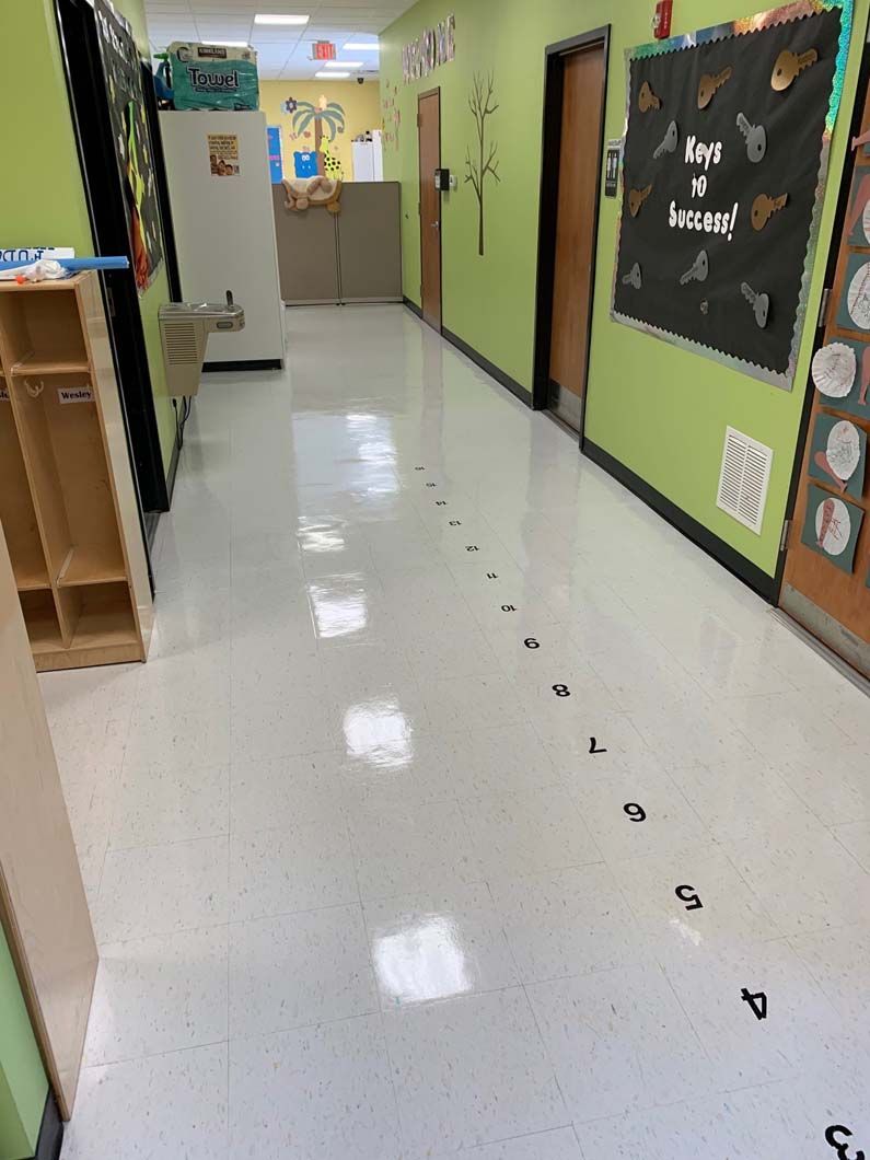 A school hallway with light green walls features a numbered floor path leading toward a classroom door.