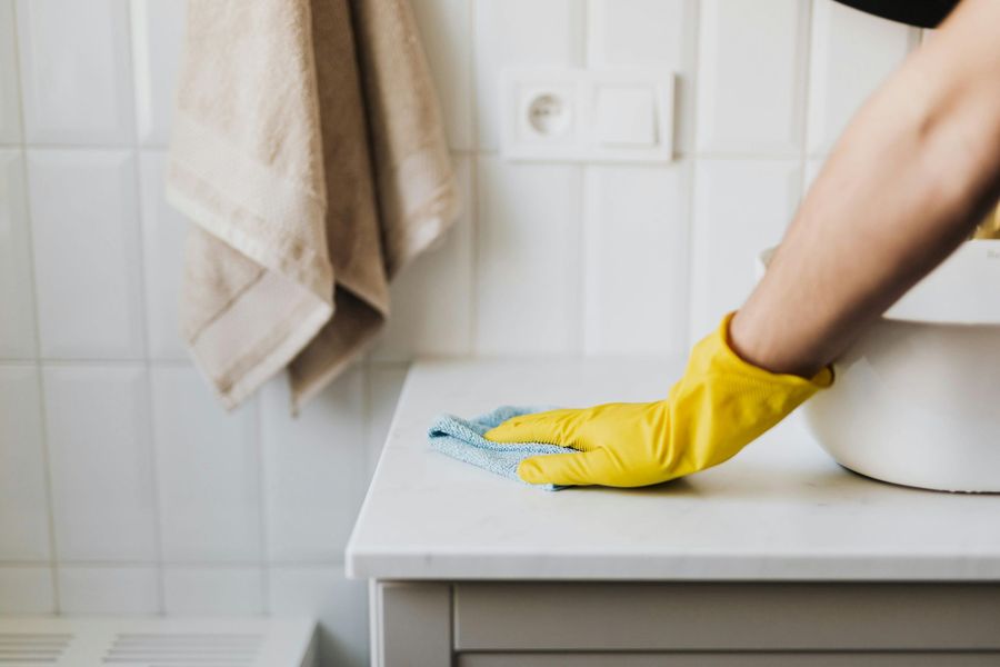 Person wearing yellow gloves cleaning a white counter in a bathroom with a blue cloth.