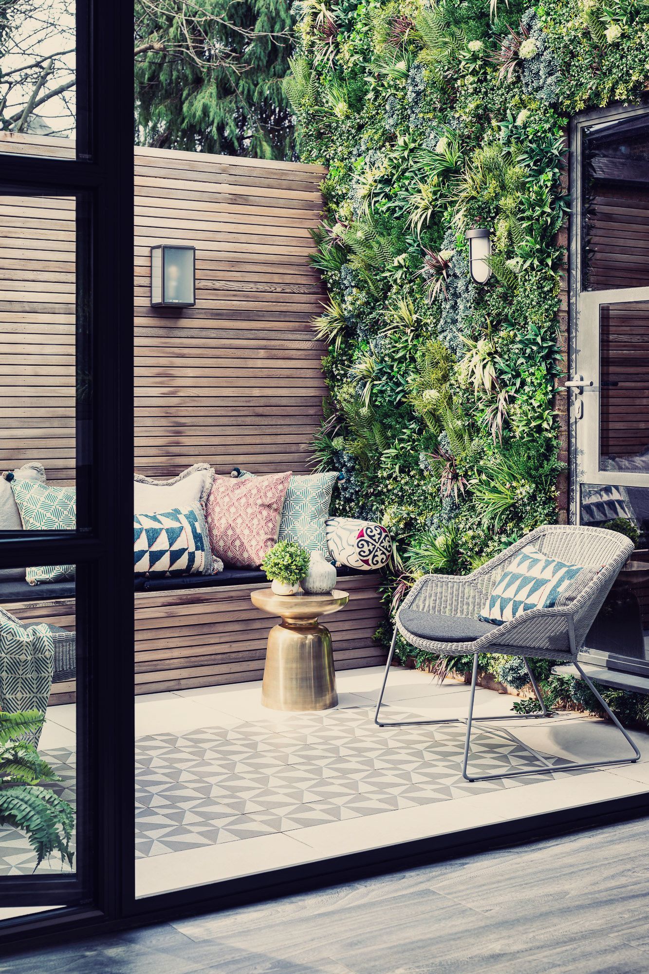 Modern patio with woven lounge chair, small table, and lush green wall viewed through a glass door