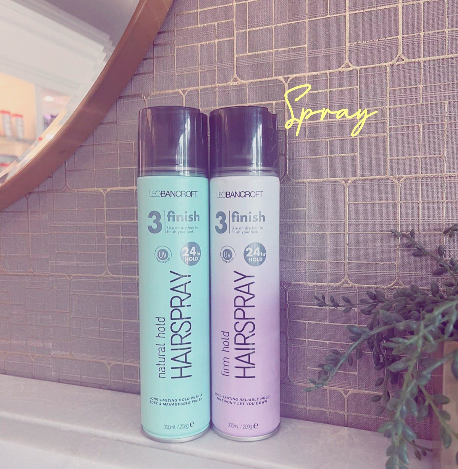 Two Pantene Hairspray cans, one teal and one lavender, standing on a shelf beside a mirror.