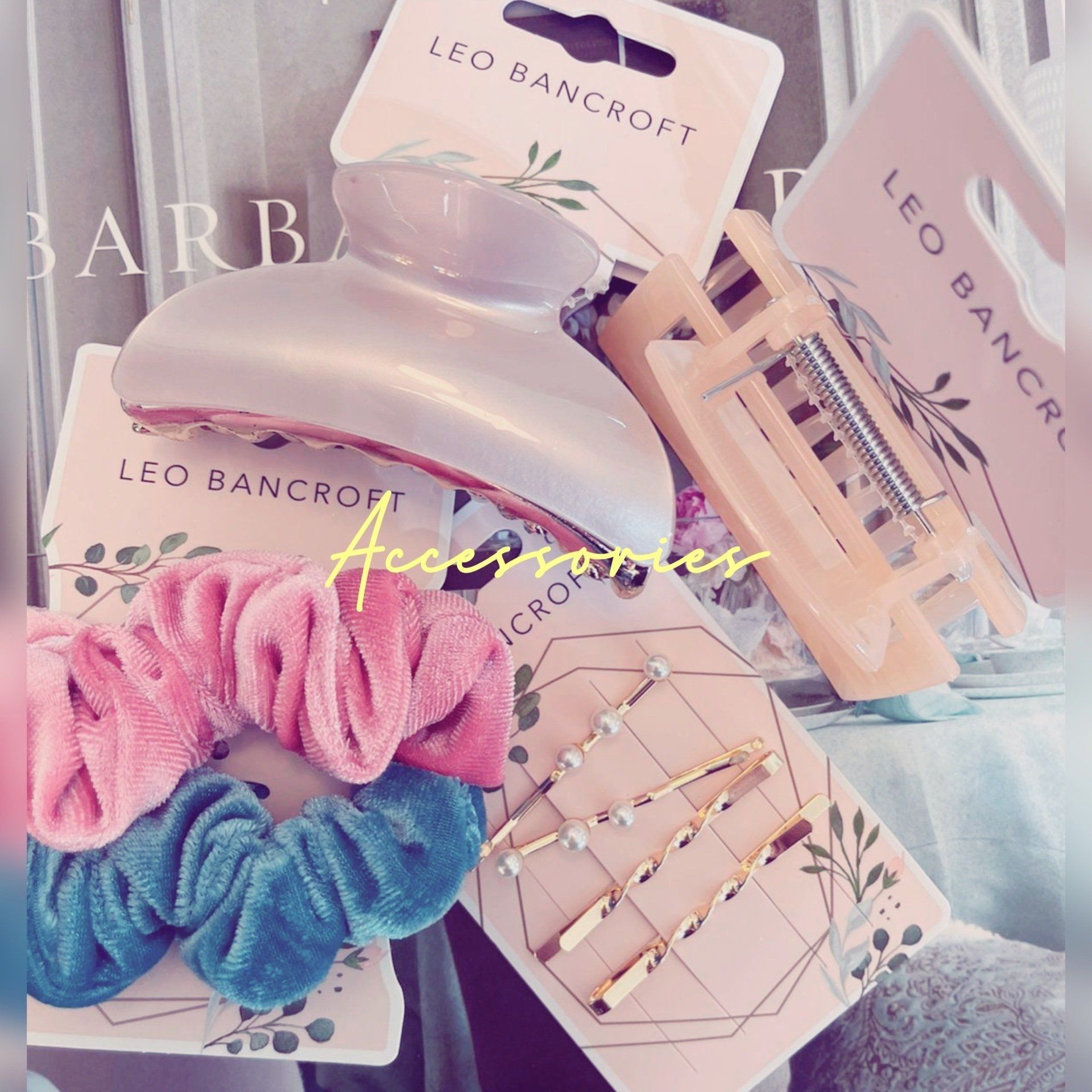 Pastel Leo Bancroft haircare products with pink scrunchies and gold hair clips on a soft fabric background