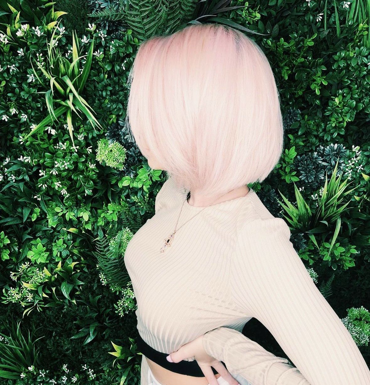 Leo Bancroft model with pastel pink bob haircut posing against a green leafy wall, wearing a cream outfit and black belt