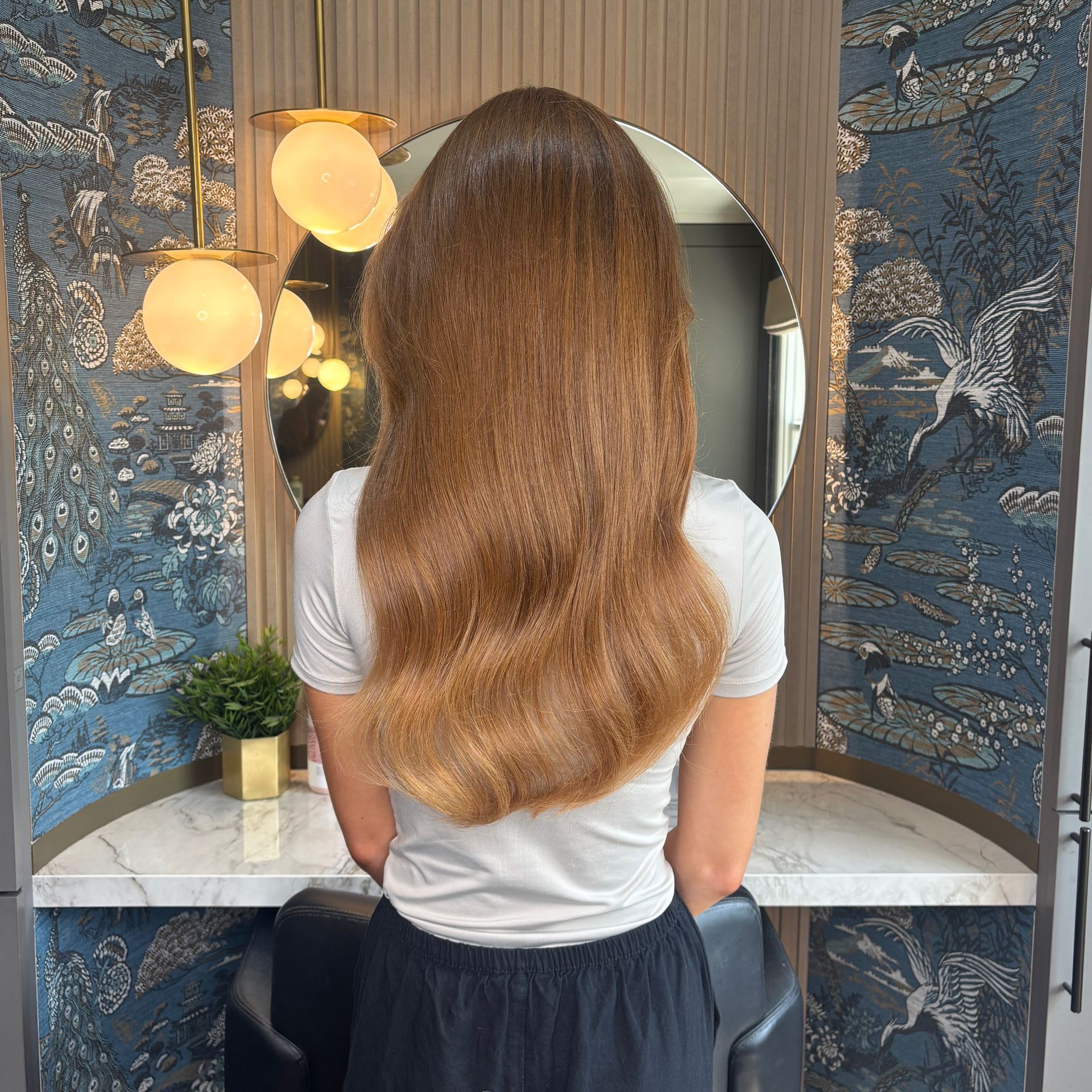 Leo Bancroft model with long wavy brown hair sitting at a vanity with a round mirror and blue patterned wallpaper.