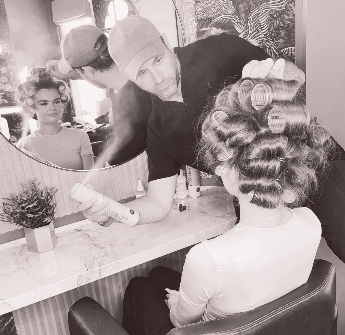 Leo Bancroft curling a client’s hair at a salon mirror, with another woman reflected in the background