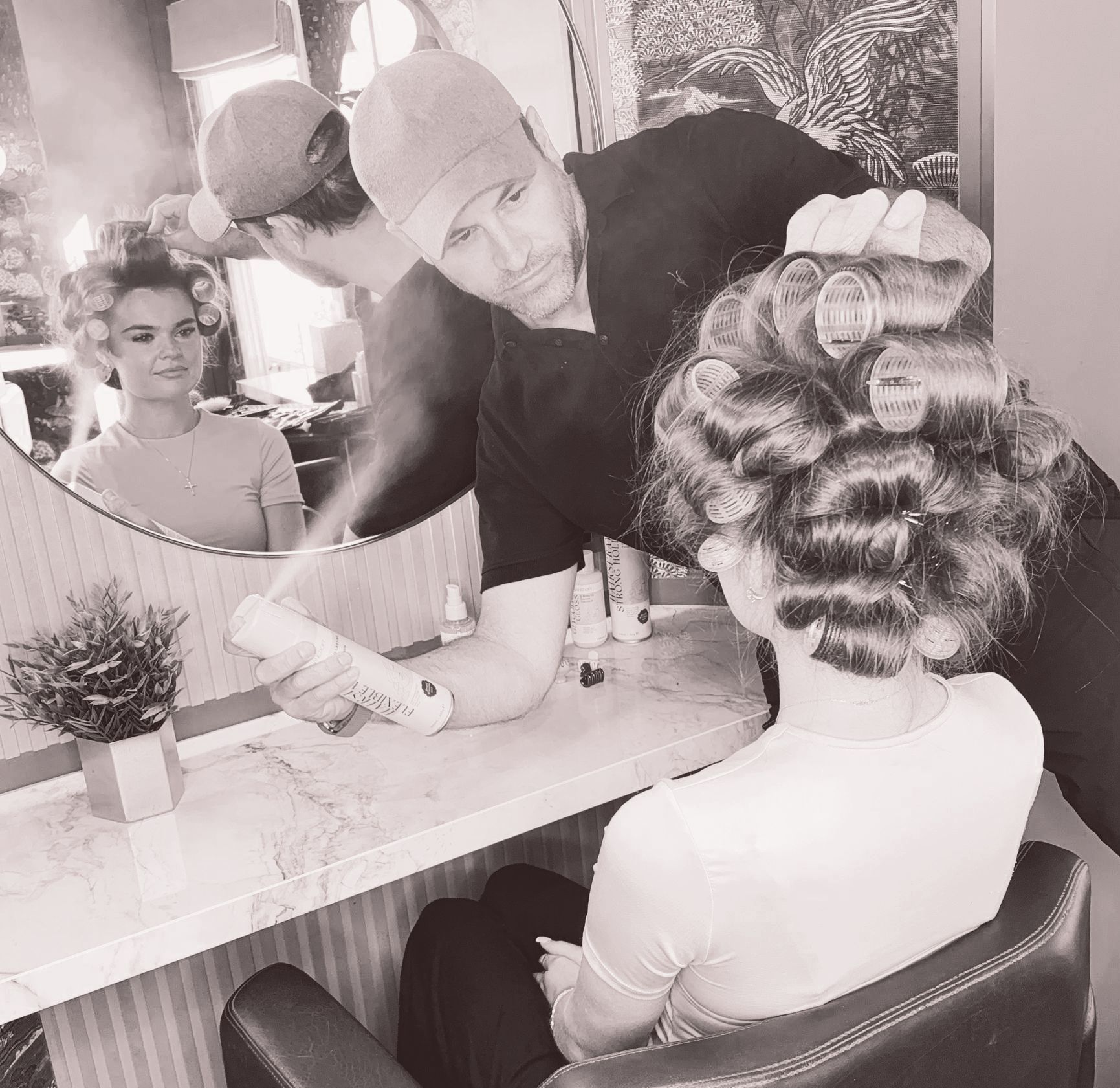Leo Bancroft curling a client’s hair at a salon mirror, with another woman reflected in the background