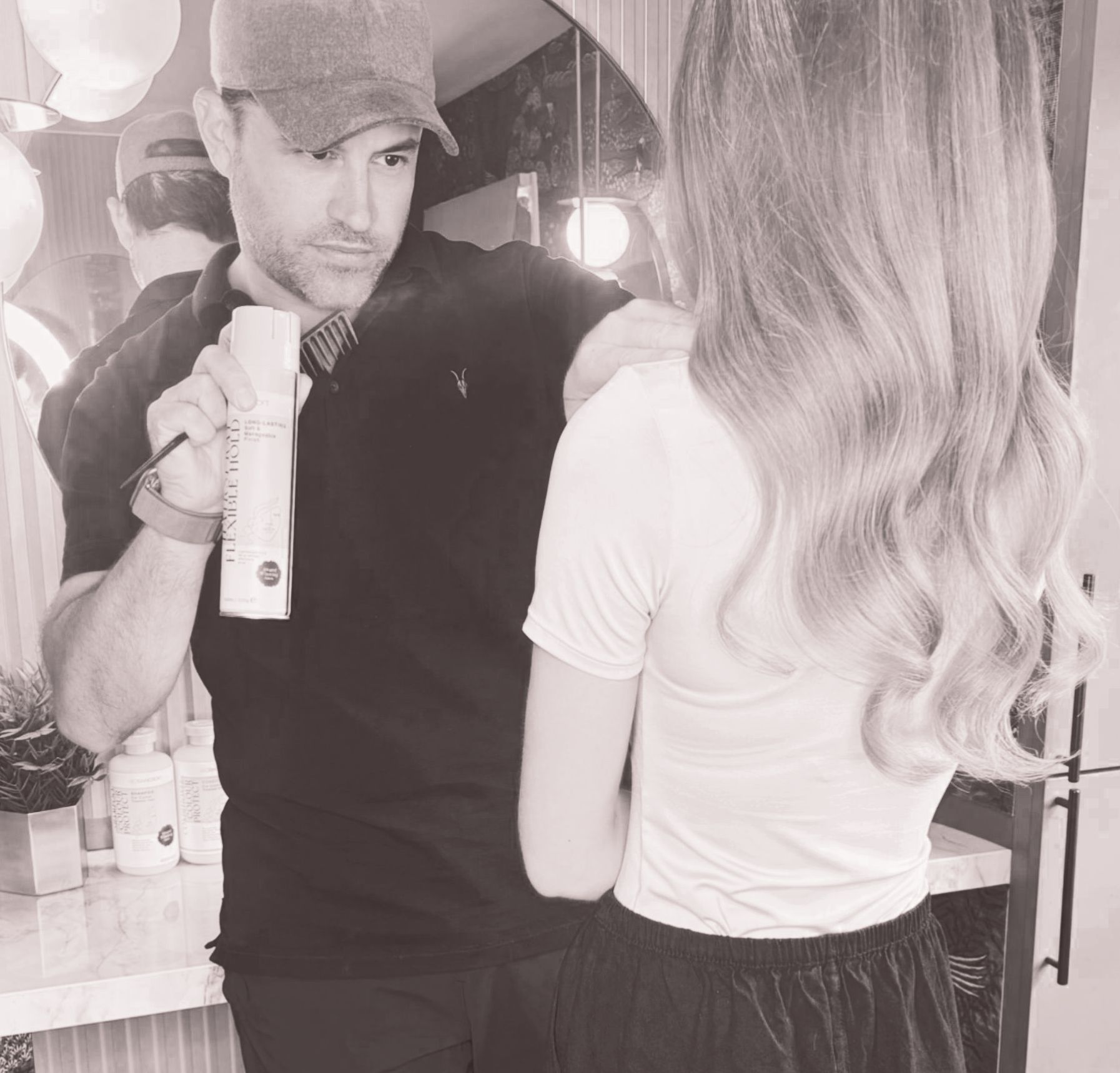 Man in black shirt and cap holding a bottle, facing a woman with long hair in a salon setting.