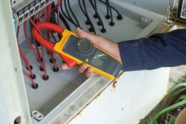 Hand holding a yellow clamp meter near electrical wiring in an open panel box