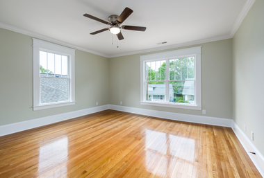 Empty bright room with hardwood floors, gray walls, white trim, and two windows with a ceiling fan.