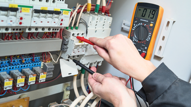 Technician testing an electrical panel with a multimeter and probe leads.