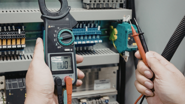Hands testing electrical wiring in a breaker panel with a multimeter and probe in a cabinet.