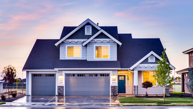 Modern two-story suburban house with a two-car garage, lit windows, and a landscaped front yard at dusk