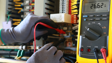 Gloved hands testing wiring with a multimeter in an electronics workspace
