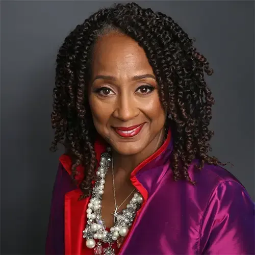 The woman is wearing a purple jacket and a pearl necklace.