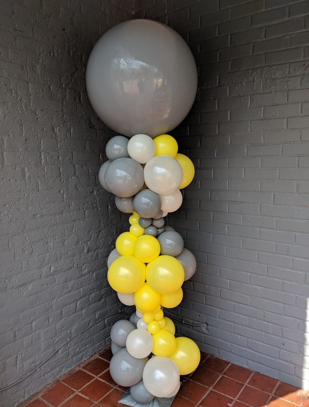 Balloon column with gray, yellow, and white balloons, topped with a large gray sphere, set against a gray brick wall.