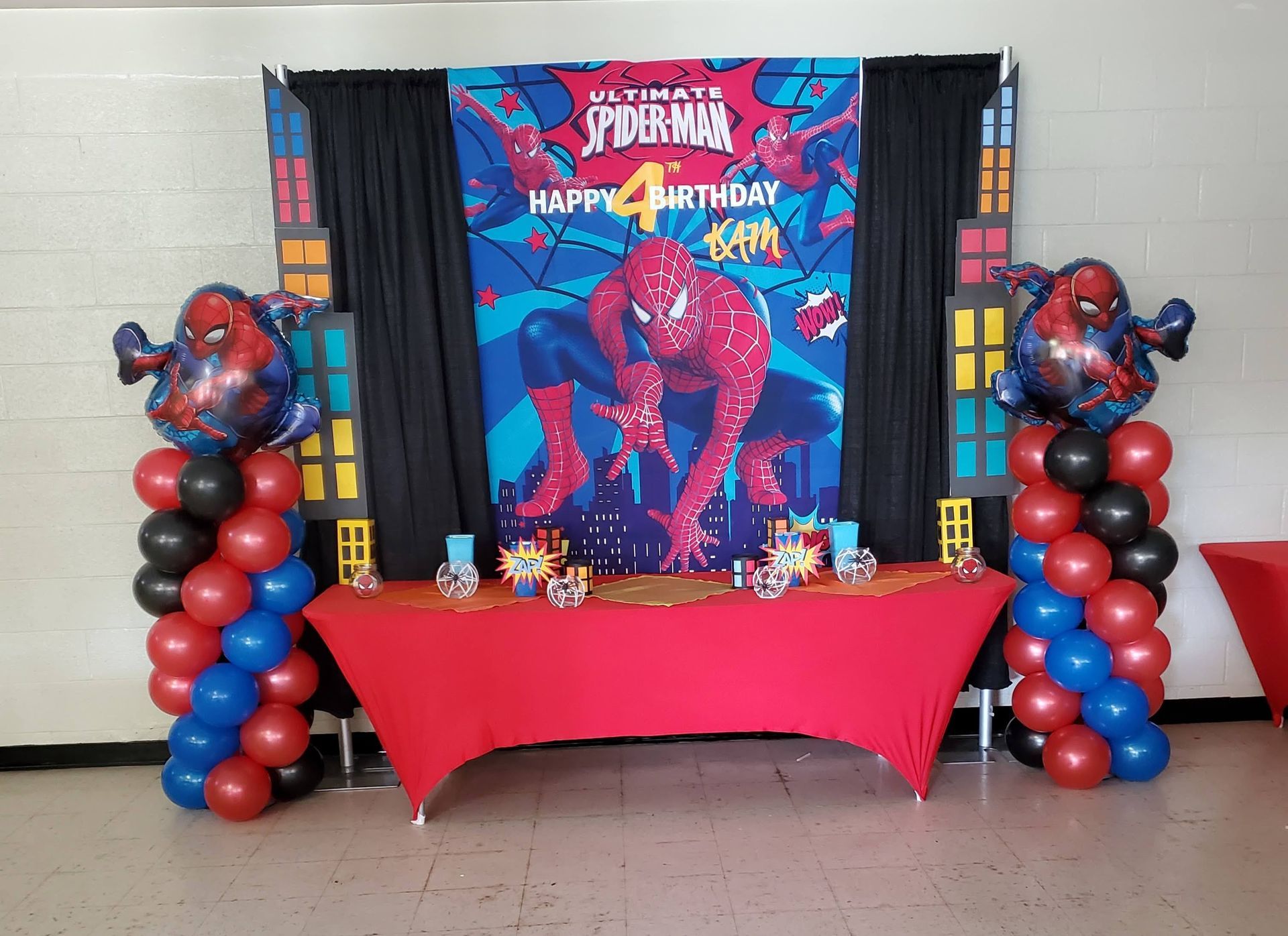 Spider-Man themed birthday party setup with a banner, red table, and balloon columns topped with Spider-Man balloons.
