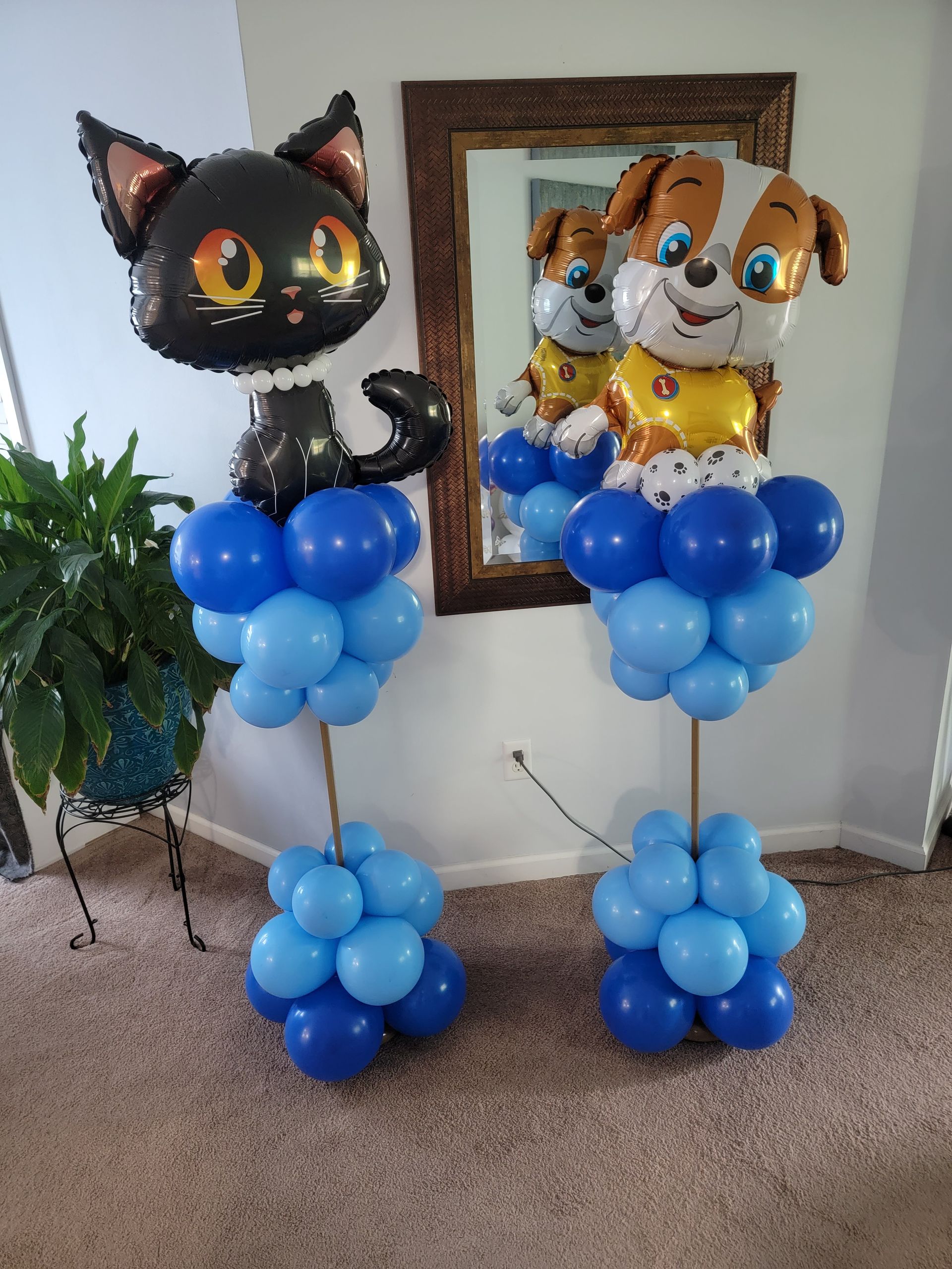 Two balloon centerpieces featuring a black cat and a dog character on stands of blue balloons in a carpeted room.