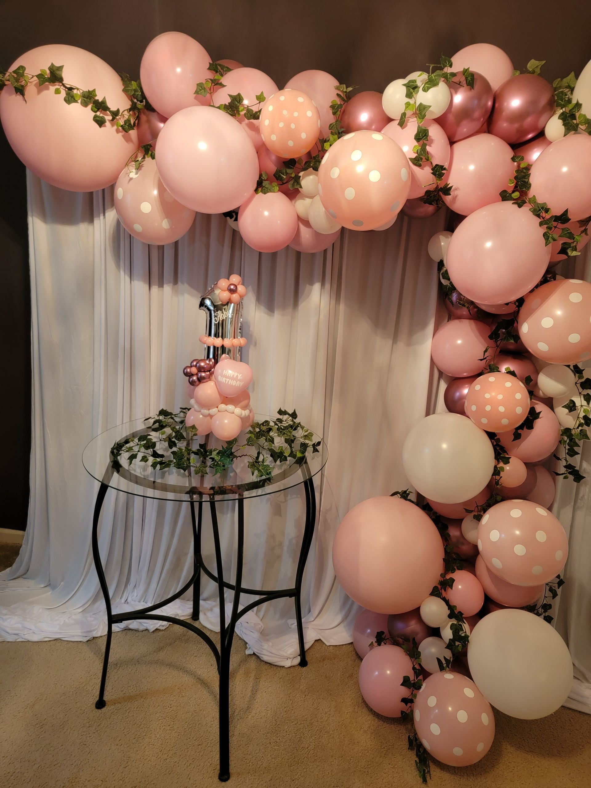 A pink and white balloon arch with greenery accents over a glass table featuring a balloon bouquet centerpiece.