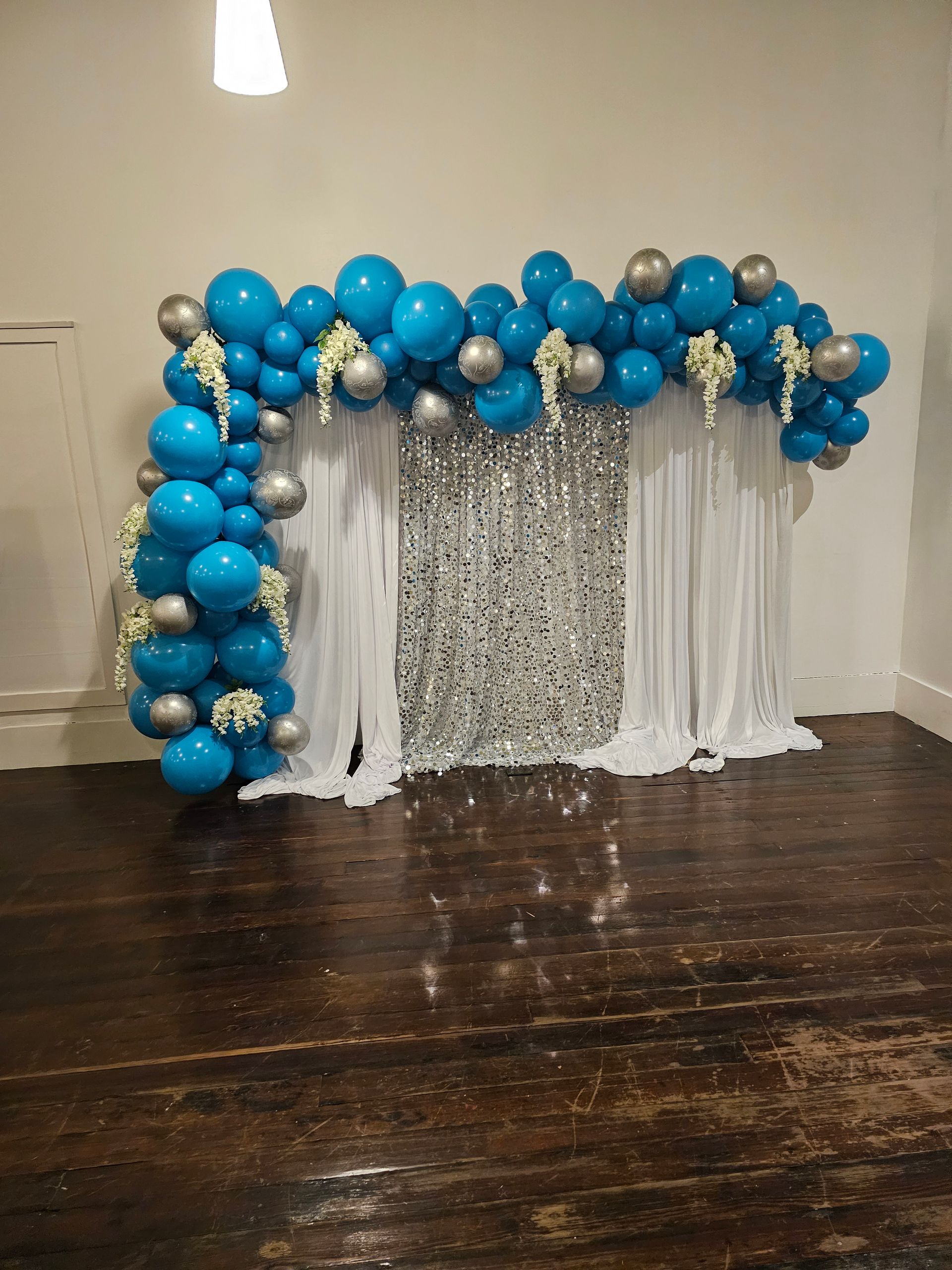 A balloon arch in blue and silver, featuring floral accents, framing a silver sequin and white fabric backdrop indoors.