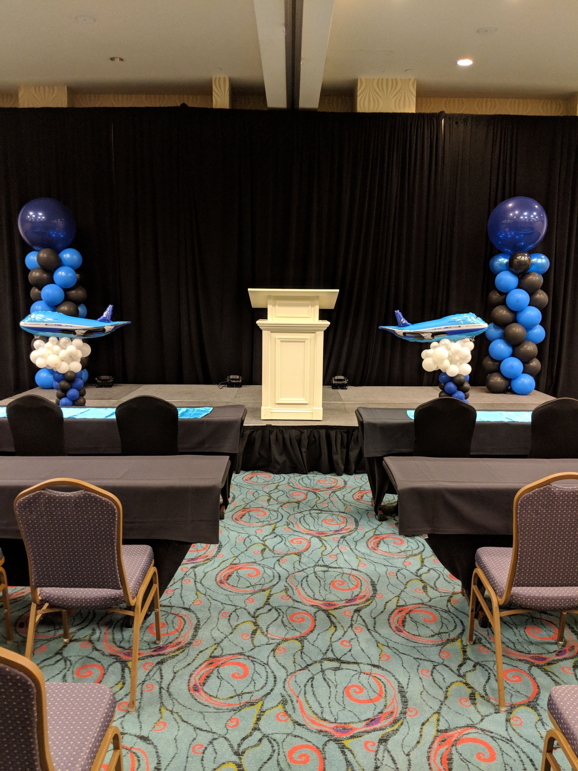 A stage with a central podium, flanked by two balloon columns topped with airplane balloons, set before a black backdrop.