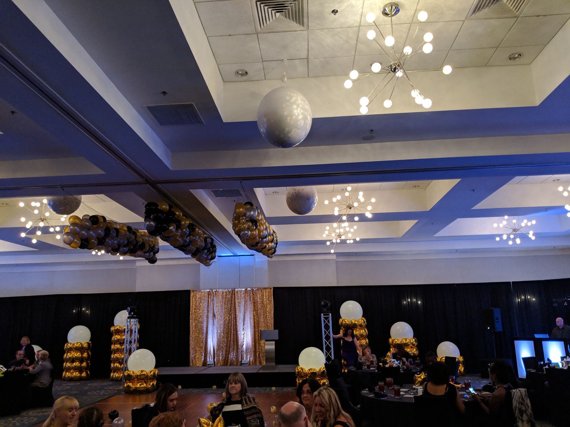 A dimly lit ballroom decorated with gold and white balloon clusters and gold curtains, set up for a formal event.