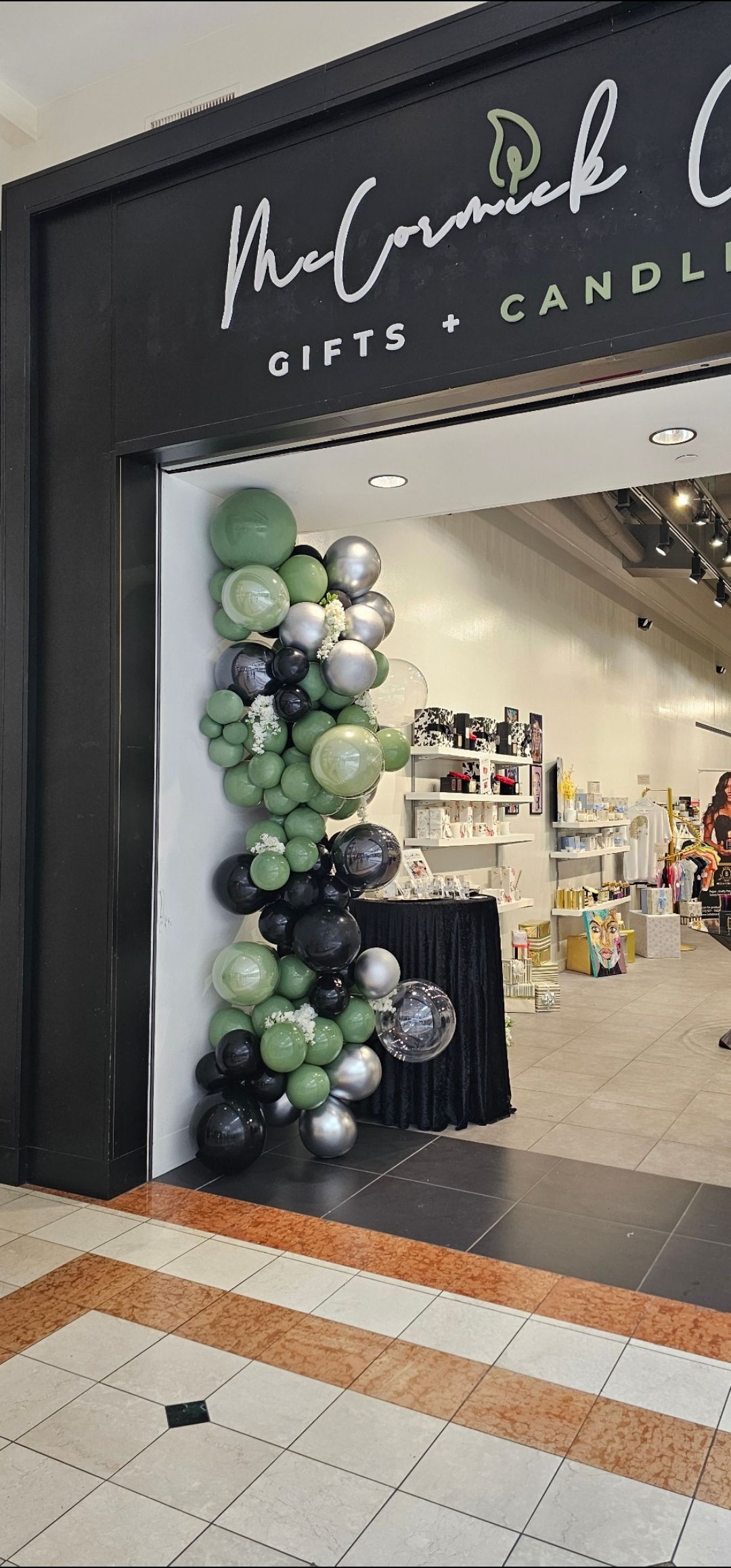 A storefront entrance decorated with a vertical balloon garland in shades of sage green, black, and silver.