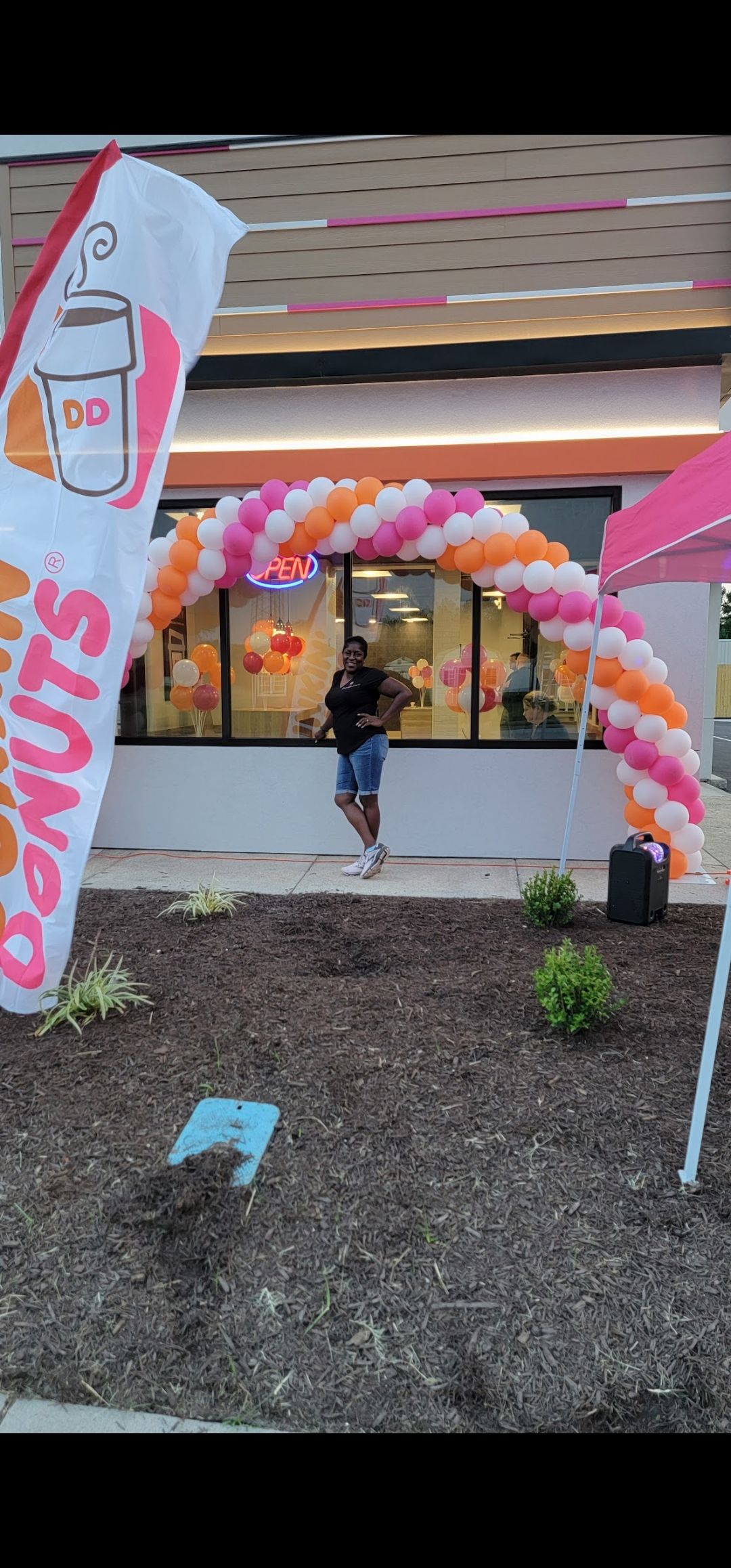 A person stands in front of a Dunkin' shop entrance decorated with a pink and orange balloon arch under a sunny sky.